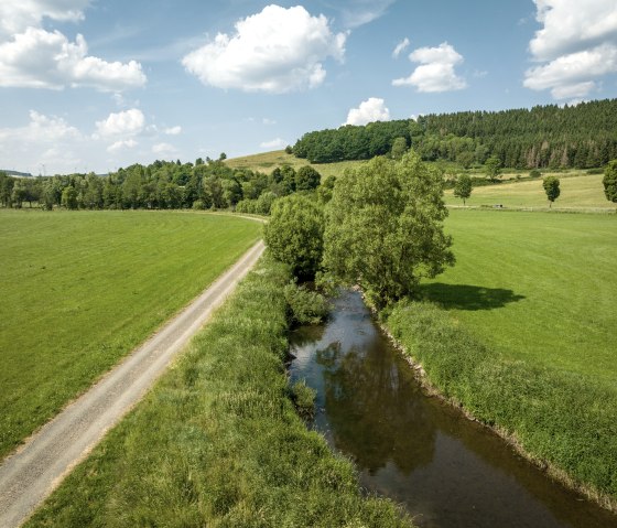 Une rivière étroite coule à travers des prairies vertes, à côté d'un chemin de terre. En arrière-plan, des collines et des arbres sous un ciel bleu avec des nuages., © Eifel Tourismus GmbH, Dominik Ketz Une rivière étroite coule à travers des prairies vertes, à côté d'un chemin de terre. En arrière-plan, des collines et des arbres sous un ciel bleu avec des nuages., © Eifel Tourismus GmbH, Dominik Ketz