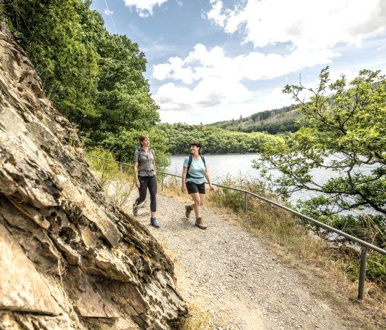 Deux femmes marchent sur un sentier étroit le long d'un lac, entouré de verdure luxuriante et de rochers. Le ciel est en partie nuageux., © Städteregion Aachen, Dominik Ketz Deux femmes marchent sur un sentier étroit le long d'un lac, entouré de verdure luxuriante et de rochers. Le ciel est en partie nuageux., © Städteregion Aachen, Dominik Ketz