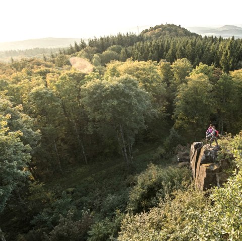 Zwei Personen stehen auf einem Felsen, umgeben von einem dichten Wald. Im Hintergrund sind Hügel und ein weiter Blick über die Landschaft zu sehen., © Eifel Tourismus GmbH, D. Ketz Zwei Personen stehen auf einem Felsen, umgeben von einem dichten Wald. Im Hintergrund sind Hügel und ein weiter Blick über die Landschaft zu sehen., © Eifel Tourismus GmbH, D. Ketz