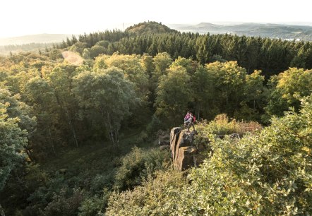 Zwei Personen stehen auf einem Felsen, umgeben von einem dichten Wald. Im Hintergrund sind Hügel und ein weiter Blick über die Landschaft zu sehen., © Eifel Tourismus GmbH, D. Ketz Zwei Personen stehen auf einem Felsen, umgeben von einem dichten Wald. Im Hintergrund sind Hügel und ein weiter Blick über die Landschaft zu sehen., © Eifel Tourismus GmbH, D. Ketz
