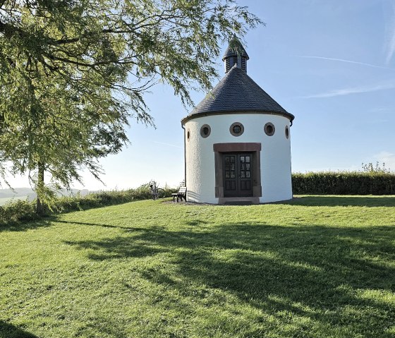 Chapelle ronde sur une pelouse verte, entourée d'arbres et sous un ciel bleu. Un vélo est adossé à la chapelle., © Touristik GmbH Gerolsteiner Land Chapelle ronde sur une pelouse verte, entourée d'arbres et sous un ciel bleu. Un vélo est adossé à la chapelle., © Touristik GmbH Gerolsteiner Land