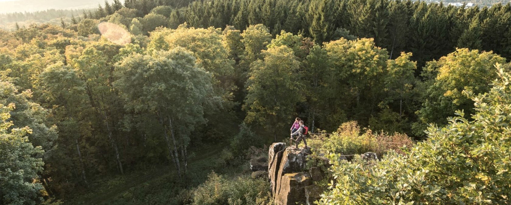 Zwei Personen stehen auf einem Felsen, umgeben von einem dichten Wald. Im Hintergrund sind Hügel und ein weiter Blick über die Landschaft zu sehen., © Eifel Tourismus GmbH, D. Ketz Zwei Personen stehen auf einem Felsen, umgeben von einem dichten Wald. Im Hintergrund sind Hügel und ein weiter Blick über die Landschaft zu sehen., © Eifel Tourismus GmbH, D. Ketz