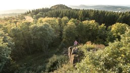 Zwei Personen stehen auf einem Felsen, umgeben von einem dichten Wald. Im Hintergrund sind Hügel und ein weiter Blick über die Landschaft zu sehen., © Eifel Tourismus GmbH, D. Ketz Zwei Personen stehen auf einem Felsen, umgeben von einem dichten Wald. Im Hintergrund sind Hügel und ein weiter Blick über die Landschaft zu sehen., © Eifel Tourismus GmbH, D. Ketz