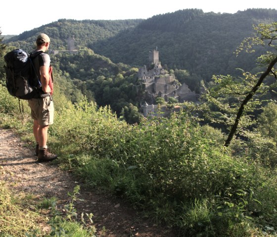A hiker with a rucksack stands on a path and looks out over the Manderscheid castles in the green hilly landscape of the Eifel., © Björn Nehrhoff A hiker with a rucksack stands on a path and looks out over the Manderscheid castles in the green hilly landscape of the Eifel., © Björn Nehrhoff
