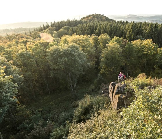 Zwei Personen stehen auf einem Felsen, umgeben von einem dichten Wald. Im Hintergrund sind Hügel und ein weiter Blick über die Landschaft zu sehen., © Eifel Tourismus GmbH, D. Ketz Zwei Personen stehen auf einem Felsen, umgeben von einem dichten Wald. Im Hintergrund sind Hügel und ein weiter Blick über die Landschaft zu sehen., © Eifel Tourismus GmbH, D. Ketz