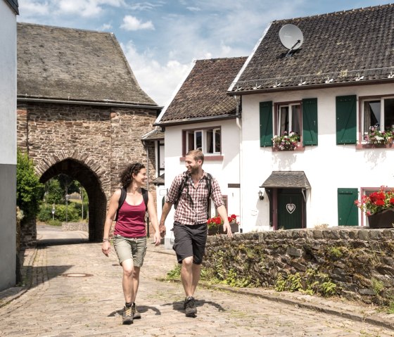 Ein Paar wandert durch den historischen Burgbering Reifferscheid, vorbei an Fachwerkhäusern und einem Torbogen., © Eifel Tourismus GmbH/D. Ketz Ein Paar wandert durch den historischen Burgbering Reifferscheid, vorbei an Fachwerkhäusern und einem Torbogen., © Eifel Tourismus GmbH/D. Ketz