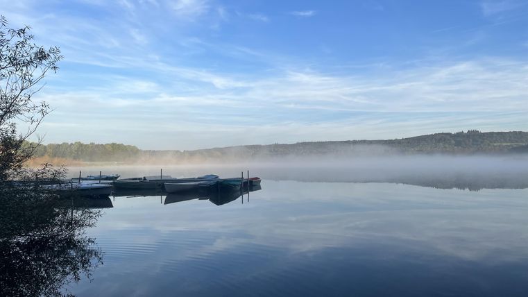 Een rustige meer met boten en een lichte mistlaag. De lucht is helder en blauw, wat een vredige sfeer uitstraalt.