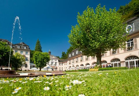Ein idyllischer Garten mit einer großen, grünen Wiese und einem Brunnen in der Mitte. Im Hintergrund befinden sich elegante Gebäude unter einem strahlend blauen Himmel.