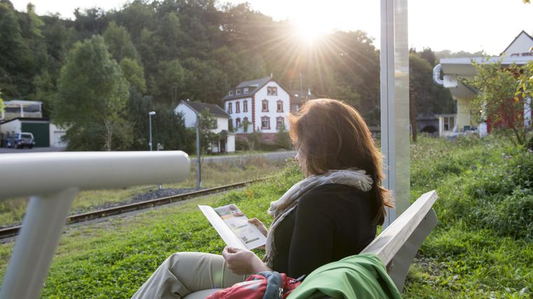 A woman is sitting on a bench and reading a magazine. In the background, the sun can be seen rising behind trees and a building.