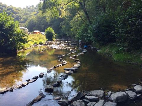 Een rustige rivier stroomt door een bosrijk gebied, met platte stenen die in het water liggen. Op de achtergrond zijn bomen en enkele tenten zichtbaar.