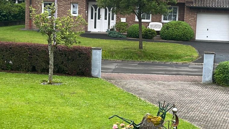 Ein gepflegtes Haus mit roter Ziegelfassade und grünem Rasen. Im Vordergrund befindet sich eine dekorative Gartenskulptur umgeben von Blumen.
