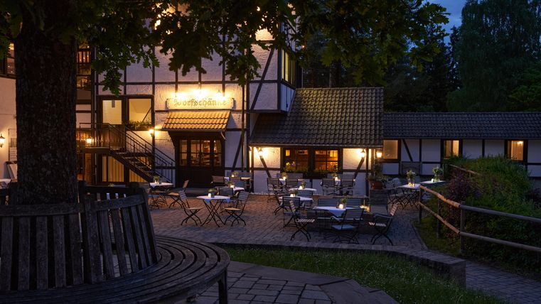 Ein charmantes Restaurant mit gemütlichen Tischen im Freien, beleuchtet bei Nacht. Die Gebäude sind im traditionellen Stil gehalten, umgeben von Bäumen und einer einladenden Atmosphäre.