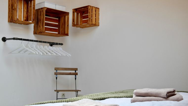 Een eenvoudige kamer met een bed en meerdere handdoeken. Aan de muur hangen houten planken en een kledingstang met kleerhangers.