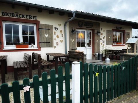 Een uitnodigend bio-bakkerijgebouw met houtsieraden en een groene omheining. Voor de ingang staan tafels en stoelen om te blijven zitten.