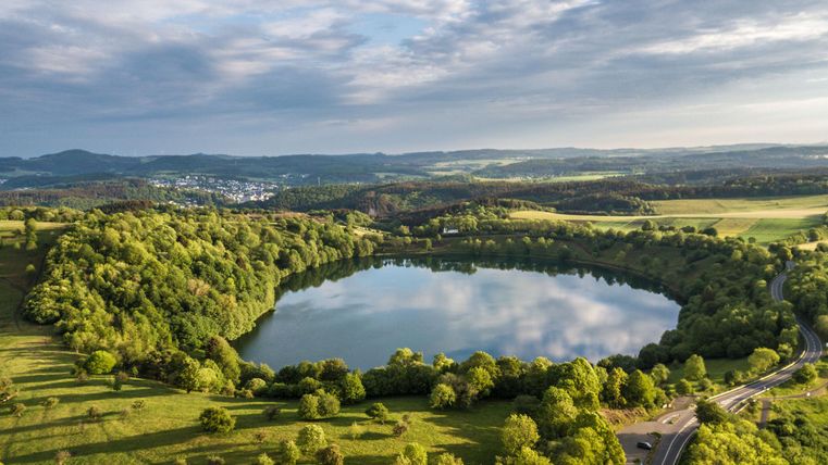 A picturesque lake surrounded by lush greenery and gentle hills. The sky is changeable with clouds and a clear blue.
