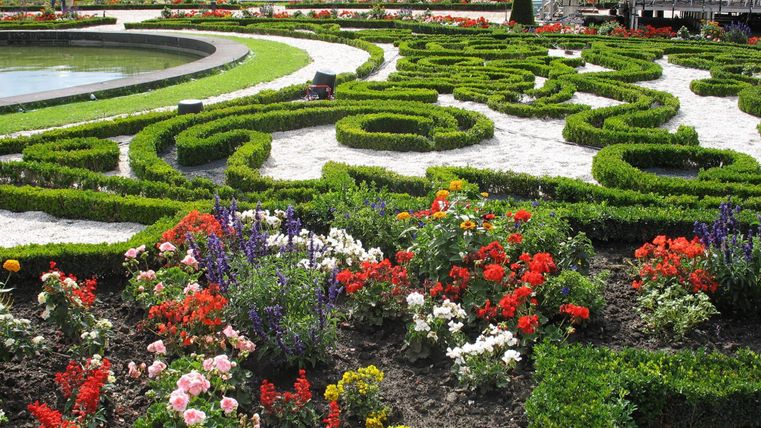 Ein schöner Garten mit kunstvoll geschnittenen Hecken und bunten Blumen. In der Mitte befindet sich ein Teich, der von grünen Pflanzen umgeben ist.