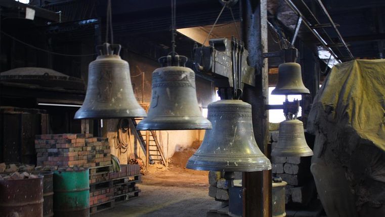 Eine Reihe von großen, gut erkennbaren Glocken hängt in einer Industriehalle. Der Raum wirkt rustikal mit Werkzeugen und Materialien im Hintergrund.