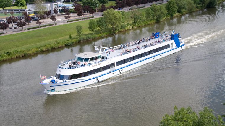 A large passenger boat is traveling on a calm river. In the background, green trees and a path can be seen.