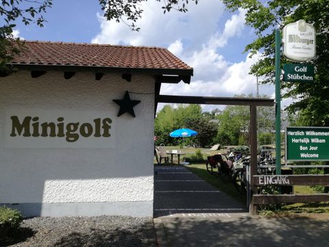 Eine Minigolf-Anlage mit einem Eingangsbereich und einem Willkommensschild. Im Hintergrund sind Bäume und ein blauer Sonnenschirm sichtbar.