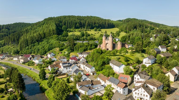 Luchtfoto van de Bertradaburg in Mürlenbach, omgeven door groene heuvels en een klein dorp. Een rivier stroomt door het landschap.