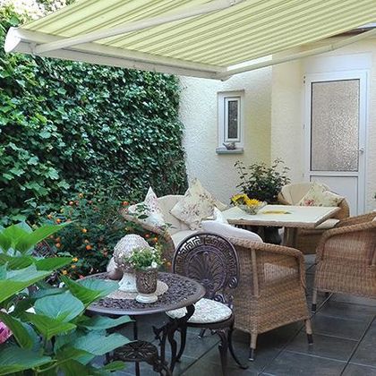 Ein gemütlicher Gartenbereich mit einem Tisch und Stühlen aus Rattan. Das Umfeld ist grün mit Pflanzen und einem Sonnenschutz.