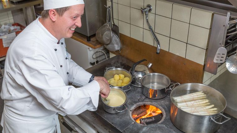 Ein Koch in einer Küche bereitet frischen Spargel und Kartoffeln zu. Er lächelt, während er am Herd arbeitet.