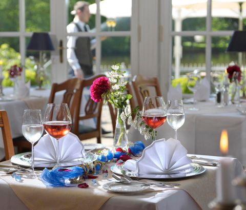 Ein elegant gedeckter Tisch in einem Restaurant mit zwei Gläsern Roséwein und dekorativen Servietten. Im Hintergrund ist ein Kellner sichtbar, während das Licht durch große Fenster strömt.