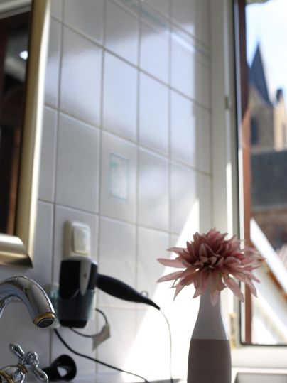 Ein heller Raum mit weißen Fliesen und einem Fenster. Auf dem Waschbecken steht eine Blumenvase mit einer pinken Blume.