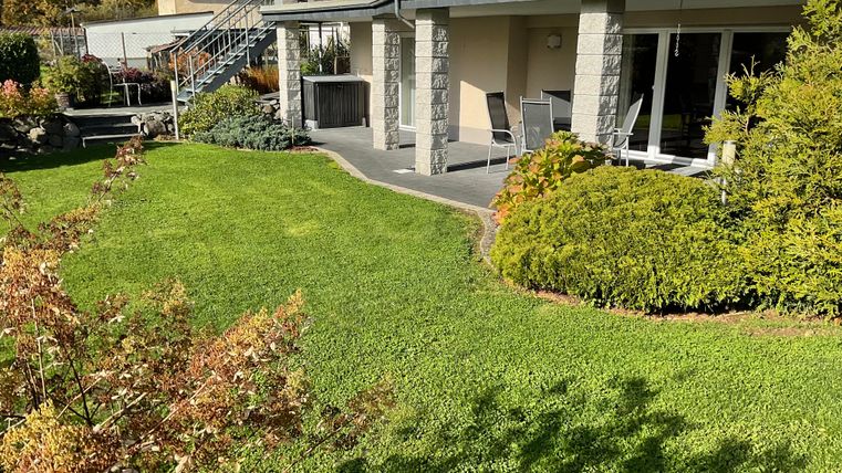 A well-kept garden with green grass and colorful shrubs surrounds a modern house. On the terrace, there are garden furniture, perfect for relaxing hours outdoors.