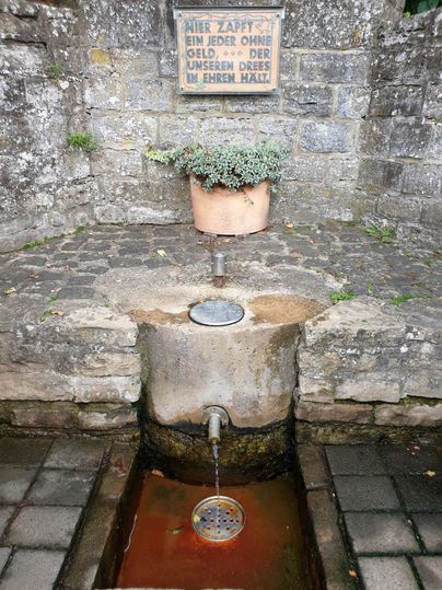 Ein kleiner Brunnen mit fließendem Wasser und einem Tontopf mit Pflanzen darüber. Die Steinmauer im Hintergrund verleiht dem Ort einen rustikalen Charme.