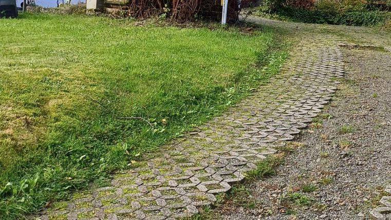 Ein gepflasterter Weg führt zu mehreren Häusern. Im Hintergrund steht ein hoher Strommast und Bäume sind sichtbar.