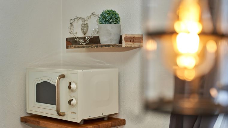 A microwave stands on a wooden shelf against the wall. Above it, plants and decorative objects are placed on another shelf.