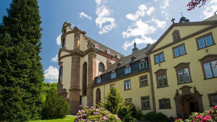 Eine historische Gebäudekombination mit einer Kirche und einem Wohnhaus. Umgeben von üppigem Grün und blühenden Blumen.