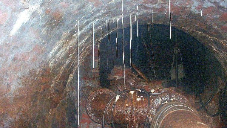 A view into a dark tunnel with red brick walls. Water droplets hang from the walls, and pipes are visible.