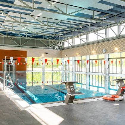 Ein modernes Hallenbad mit einem blauen Schwimmbecken und großen Fenstern. Die Umgebung ist hell und freundlich, mit bunten Fähnchen, die am Beckenrand hängen.