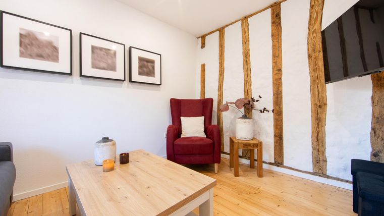 Ein gemütlicher Wohnraum mit einem roten Sessel und einem Holztisch. Weiße Wände und dekorative Bilder verleihen dem Raum eine einladende Atmosphäre.