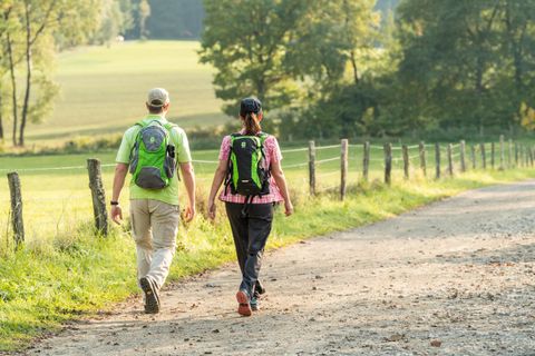 A couple is hiking on a rural path, surrounded by trees and meadows. Both are wearing backpacks and enjoying nature.