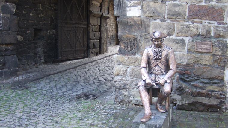 Eine Statue eines sitzenden Mannes aus Bronze, die in einer steinernen Gasse platziert ist. Im Hintergrund sind historische Mauern und ein Tor zu sehen.