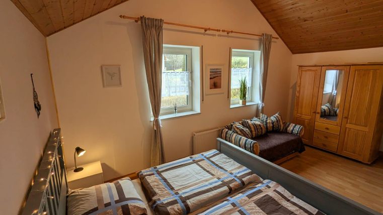 A bright bedroom with two beds and a cozy style. There are large windows, a wardrobe, and a seating area.