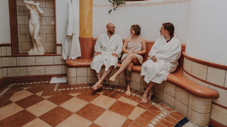 Drei Personen sitzen in einem Wellnessbereich in Bademänteln. Im Hintergrund ist eine Statue und eine gemütliche Atmosphäre zu sehen.