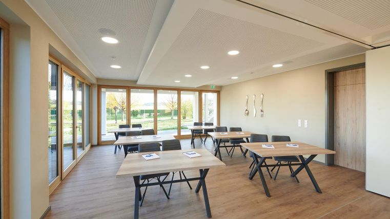 Ein moderner Besprechungsraum mit großen Fenstern und Holzböden. Der Raum ist mit Tischen und Stühlen ausgestattet, bereit für ein Treffen.