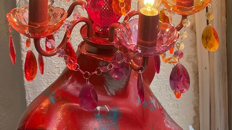 Een kleurrijke lamp in de vorm van een menselijk bovenlichaam. De lamp heeft meerdere gloeilampen en is versierd met glazen kralen.