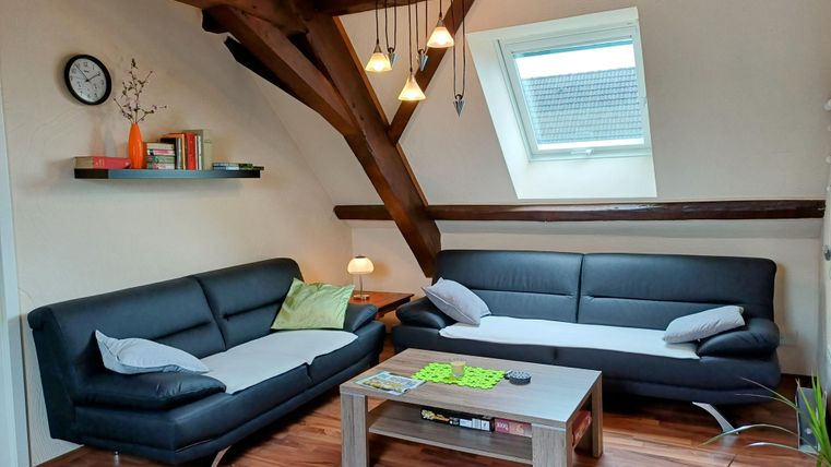 A cozy living room with two black sofas and a wooden table. Large windows let in plenty of light, and the ceiling beams give the room a special charm.