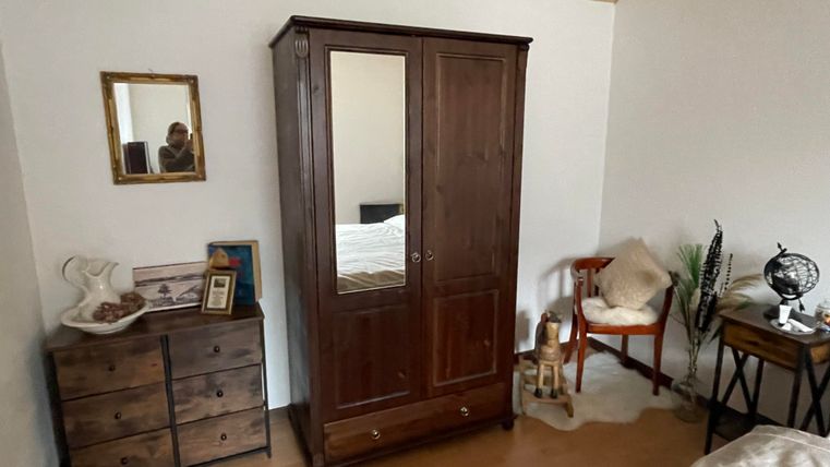 Een gezellige slaapkamer met een klassieke kast en een nachtkastje. Op de commode liggen persoonlijke spullen en een kleine plant staat naast de stoel.