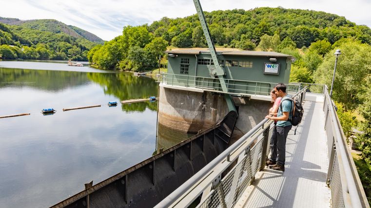 Ein Paar steht auf einer Aussichtsplattform an einem Staudamm. Im Hintergrund sind grüne Hügel und ein ruhiger See zu sehen.