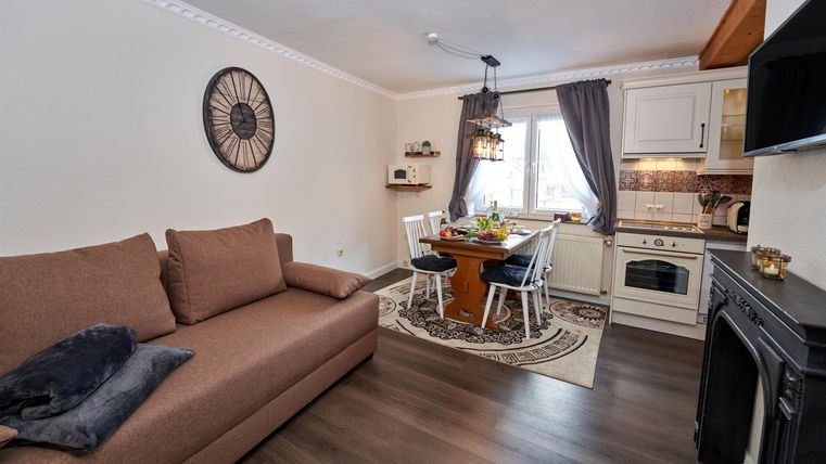 A cozy living room with a brown sofa and a dining table. The kitchen is well-equipped and the window lets in a lot of light.