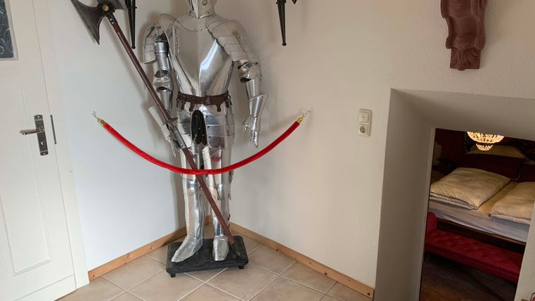 A knight in shining armor stands by the wall next to a red barrier tape. In the background, a tastefully decorated room with a bed is visible.