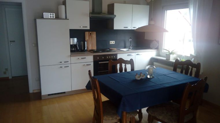 A bright kitchen with modern white furniture and a wooden table. The table is set with blue tablecloths and there are windows with plenty of natural light.