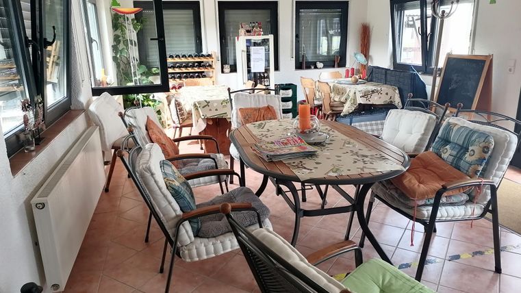 Ein gemütlicher Raum mit mehreren Sitzgelegenheiten und einem Tisch. Große Fenster lassen viel Licht herein und es gibt dekorative Elemente auf den Tischen.