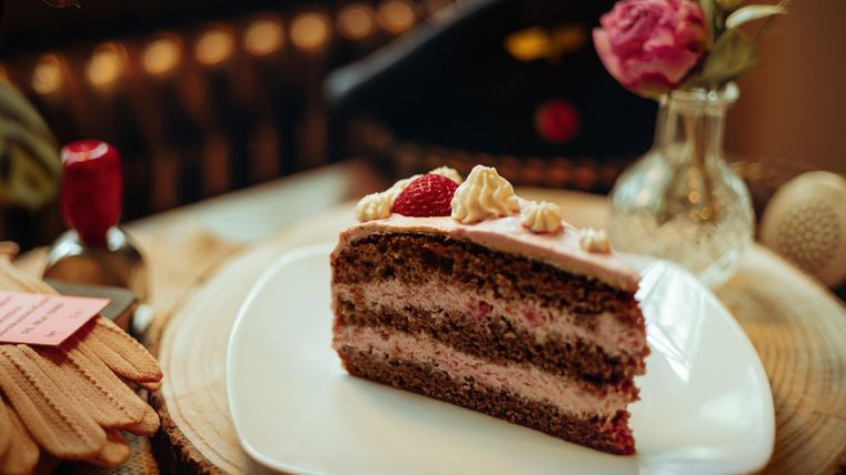 A piece of chocolate cake with heavenly filling and decorative berries. The cake is on a white plate and is accompanied by a pretty flower in the background.
